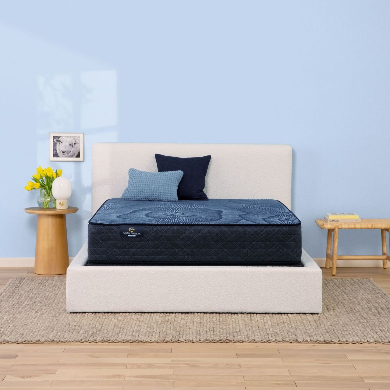 A blue mattress sits on a white bed frame in a room with a light blue wall, a wood side table, and a small wood bench.