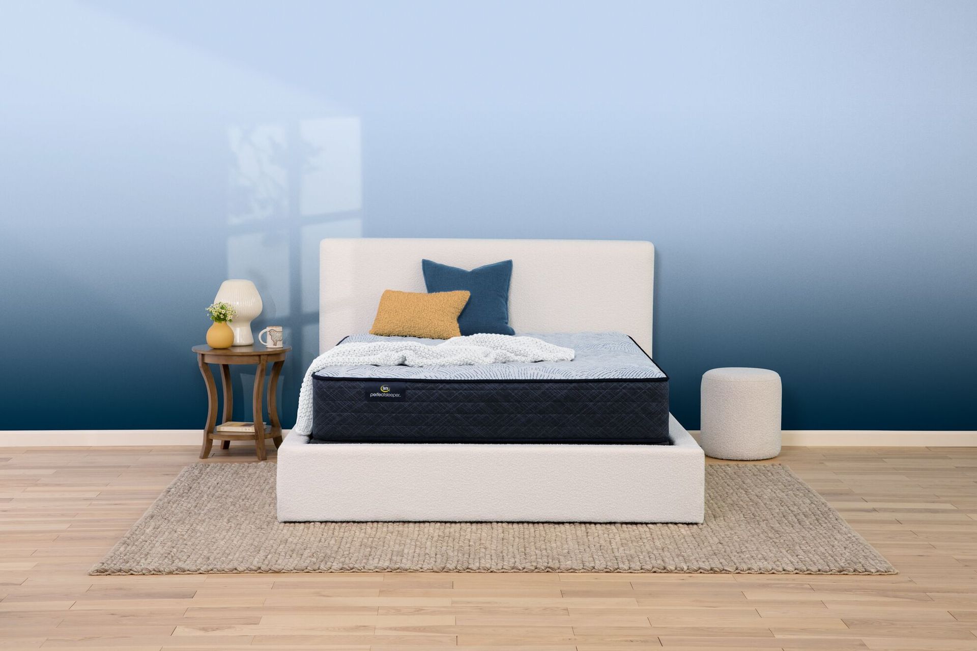 A mattress on a white platform bed frame in a room with light wood floors, a rug, a nightstand, and a blue gradient wall.