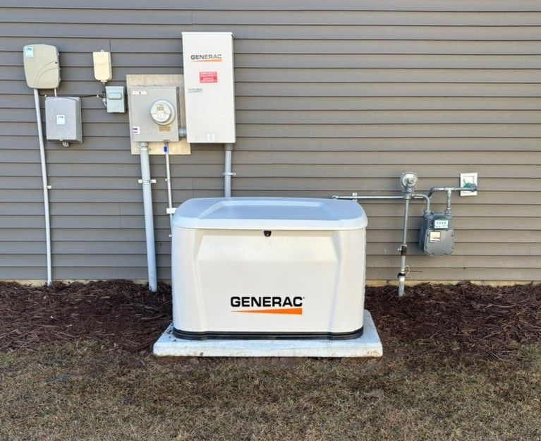 A Generac home generator sits next to a brick house with a flower bed. It is grey with a white base.