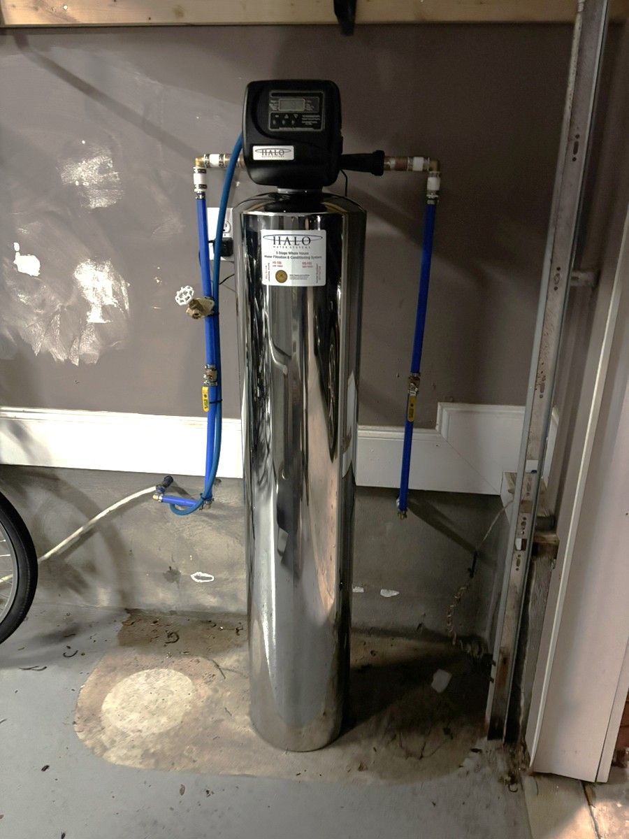 Chromed water softener tank with black control head and blue hoses in a garage-like utility area