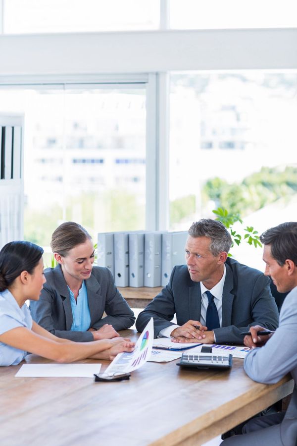Business team in suits at a meeting, reviewing documents around a table in a bright office.