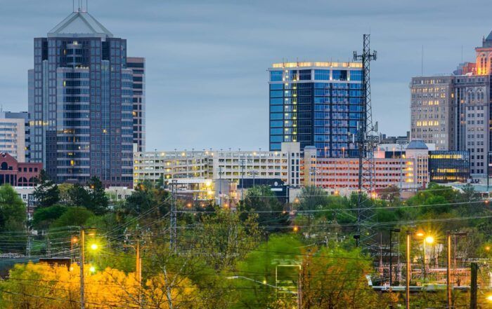 City skyline at dusk with lit buildings, trees, and streetlights.