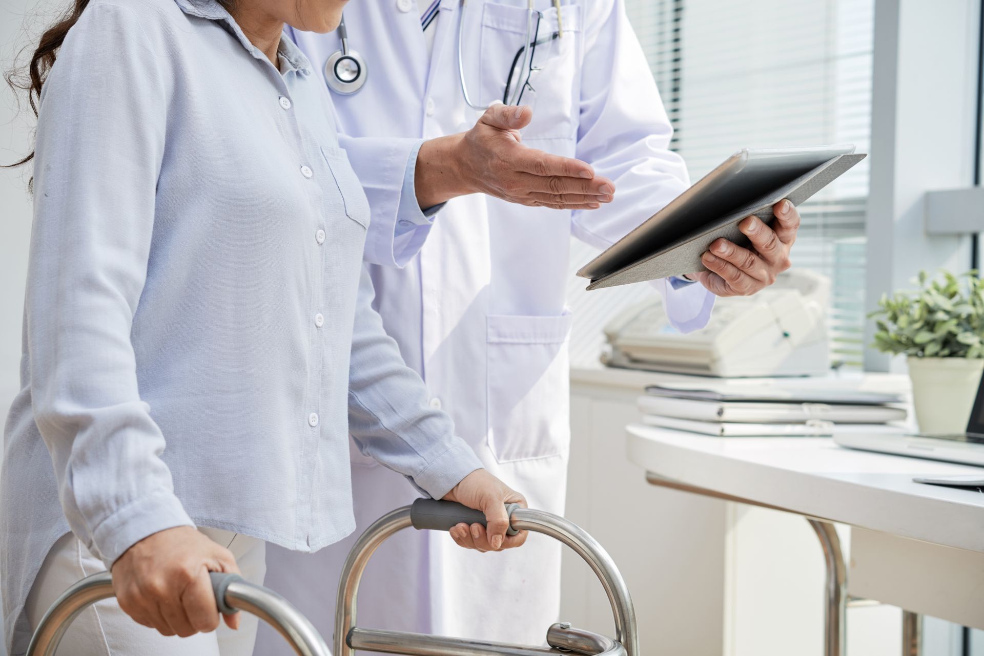 Woman using a walker consults with a doctor in an office. Doctor points to tablet.