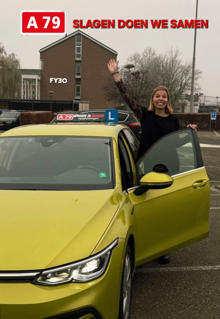 Vrouw in zwart shirt met gele auto zwaait, vierend dat ze geslaagd is voor haar rijexamen voor een gebouw.