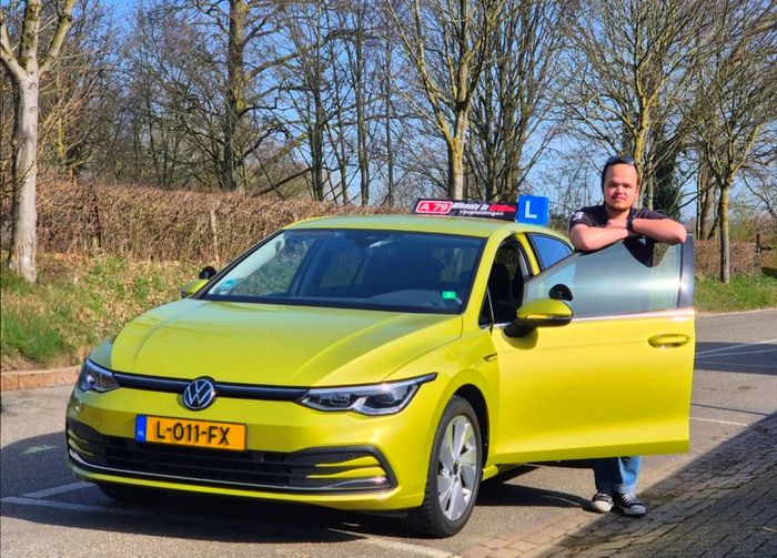 Een man staat naast een felgele Volkswagen Golf, een lesauto. Het bestuurdersportier staat open. Kentekenplaat zichtbaar.