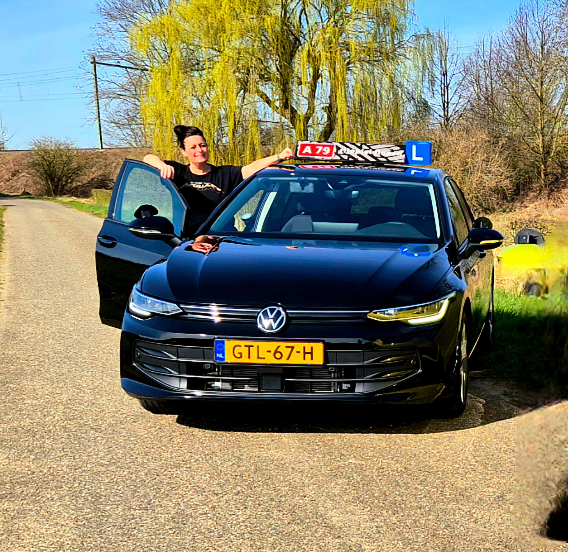 Vrouw leunt tegen een zwarte Volkswagen met een bordje voor beginnende bestuurders. Zonnige dag, landelijke omgeving.