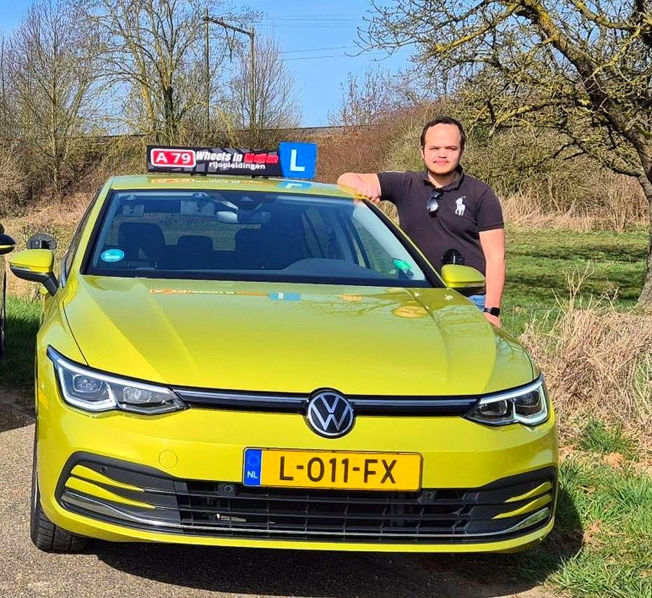 Een man staat naast een gele Volkswagen, een lesauto van de rijschool.