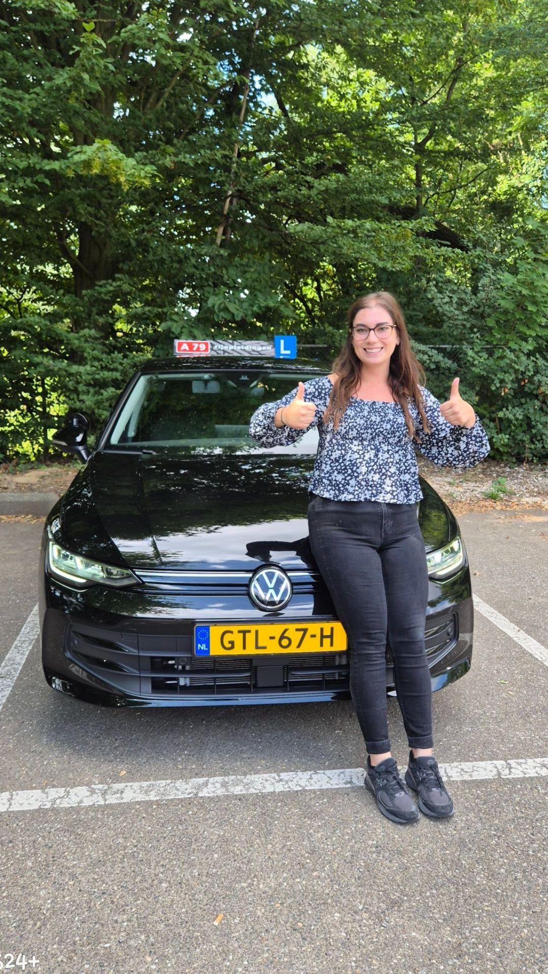 Vrouw met opgestoken duim naast een zwarte auto met kentekenplaten in een parkeerplaats.
