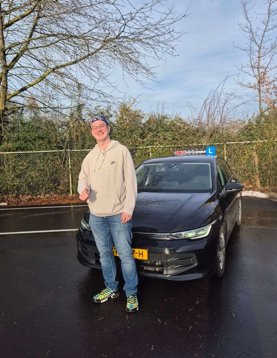 Een man in spijkerbroek en hoodie staat lachend naast een zwarte auto met een 