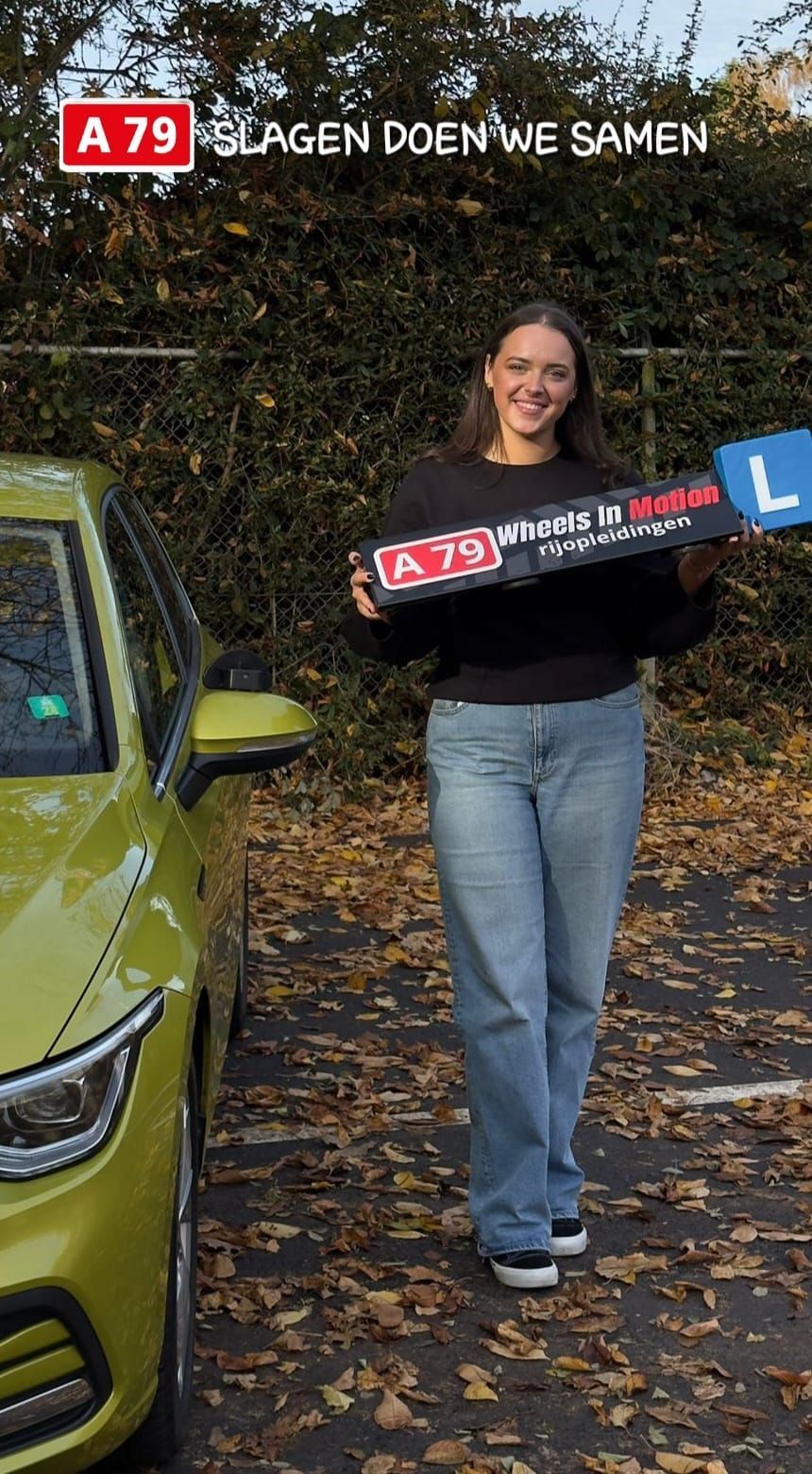 Vrouw met een bord naast een gele auto, lachend. Op het bord staat: 