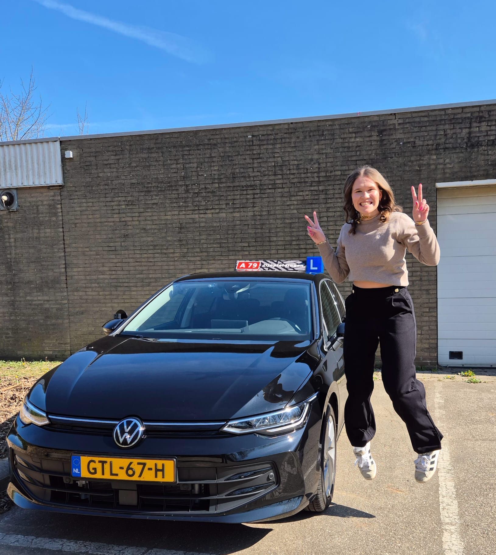 Een vrouw springt van vreugde naast een zwarte auto met een bord van een rijschool.
