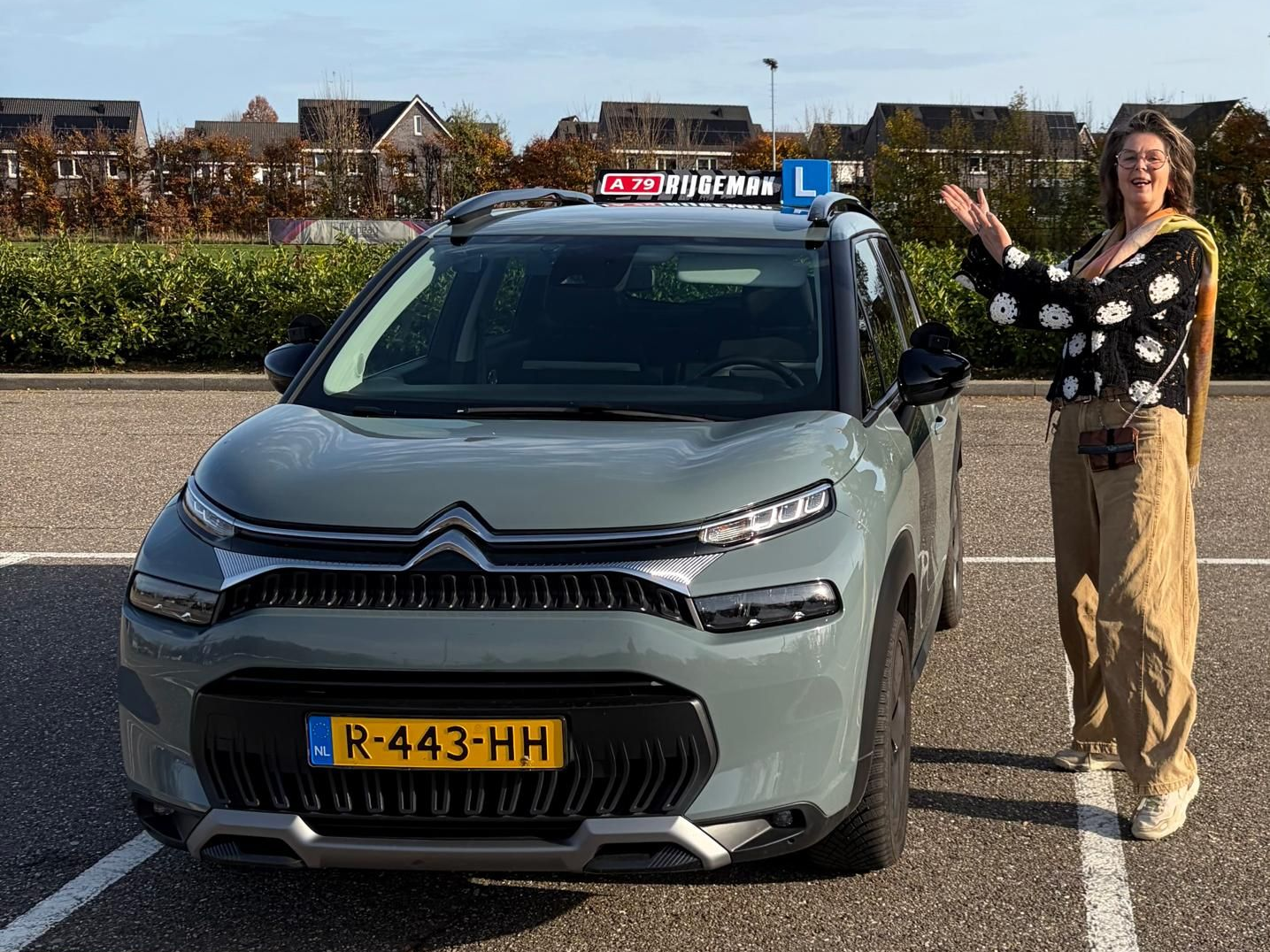 Een vrouw staat naast een grijze Citroën, waarschijnlijk een lesauto van een rijschool, met 