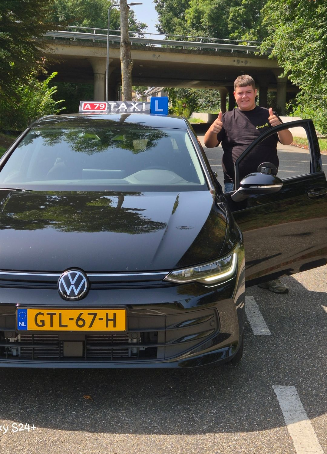 Een man steekt zijn duim omhoog naast een zwarte Volkswagen, een lesauto, onder een brug door.
