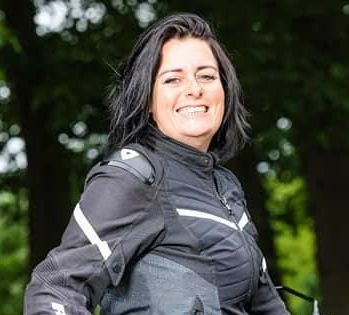 Een lachende vrouw, gekleed in een zwarte motorjas, staat buiten met bomen op de achtergrond.