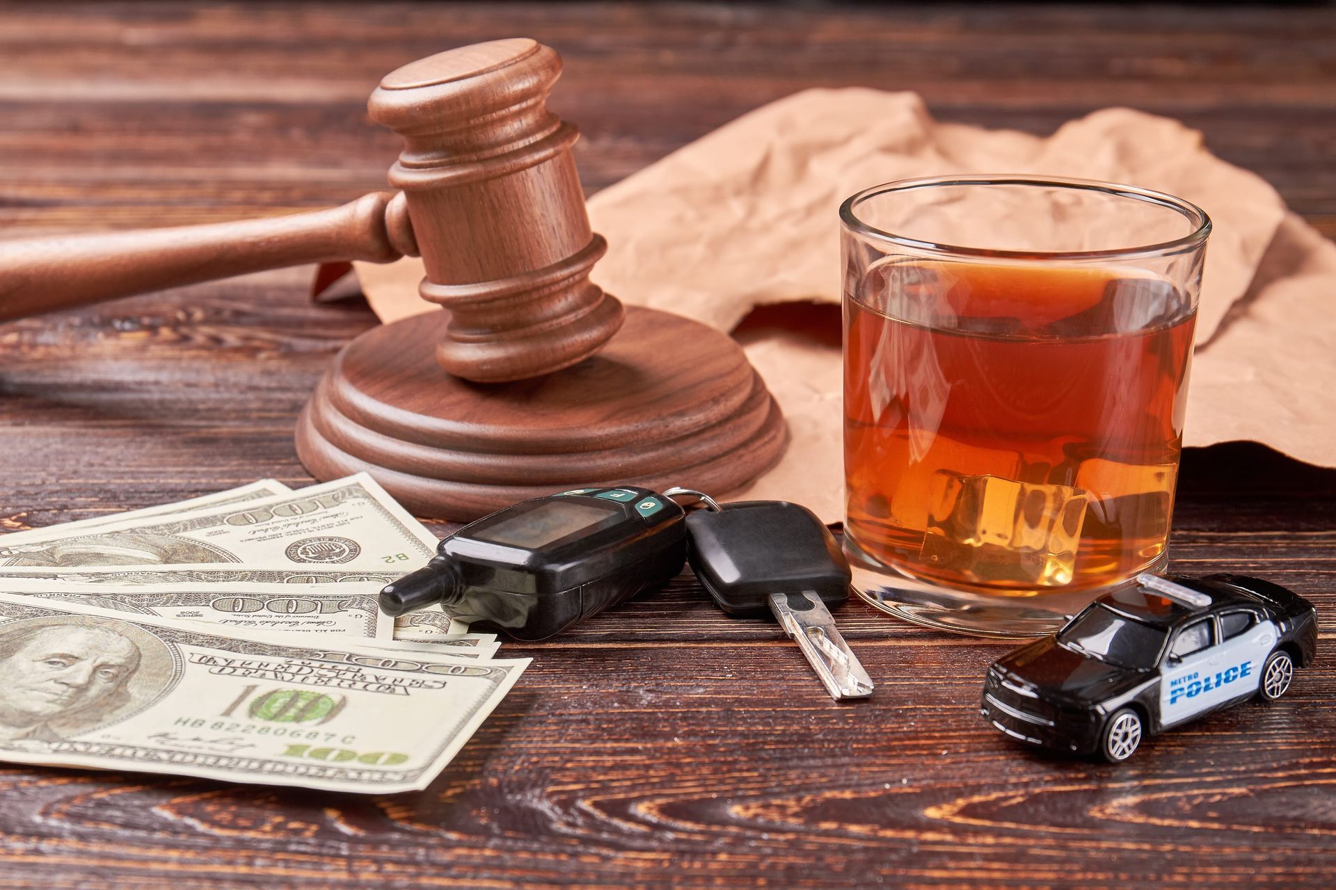 Gavel beside whiskey, car keys, and cash on a wooden table, suggesting legal or DUI-related consequences