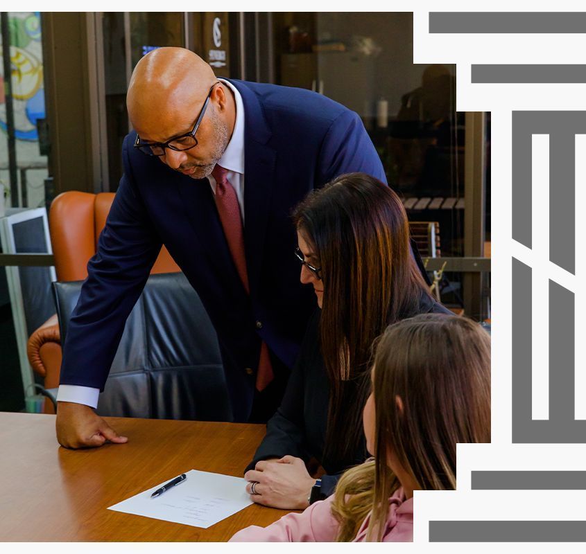 A person in a suit leans over a table to review a document with two seated individuals in an office setting.