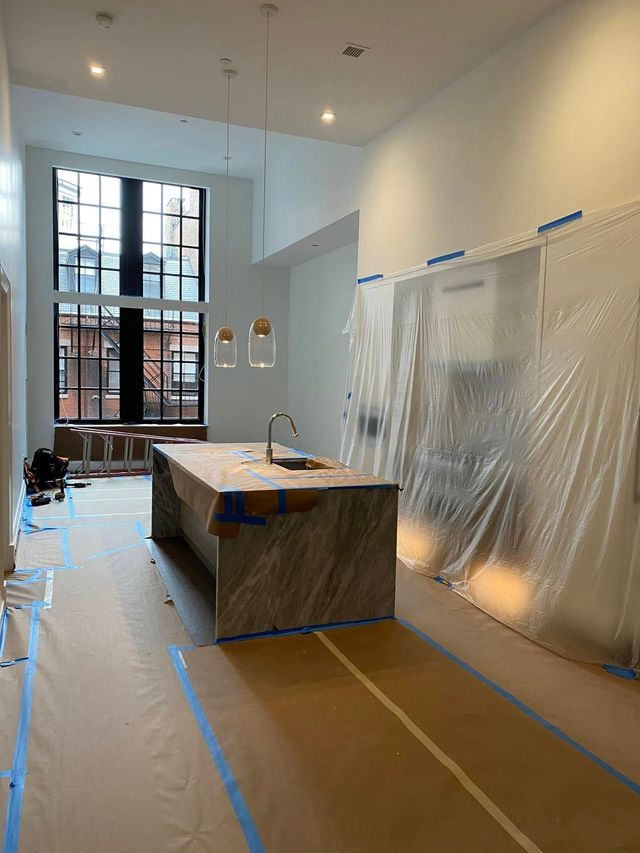 Bright kitchen under renovation with plastic-covered walls, blue tape on floor, and an island in the center.