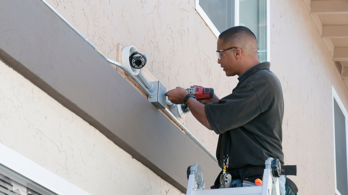 Security System Repair A man is working on a security camera on the side of a building.