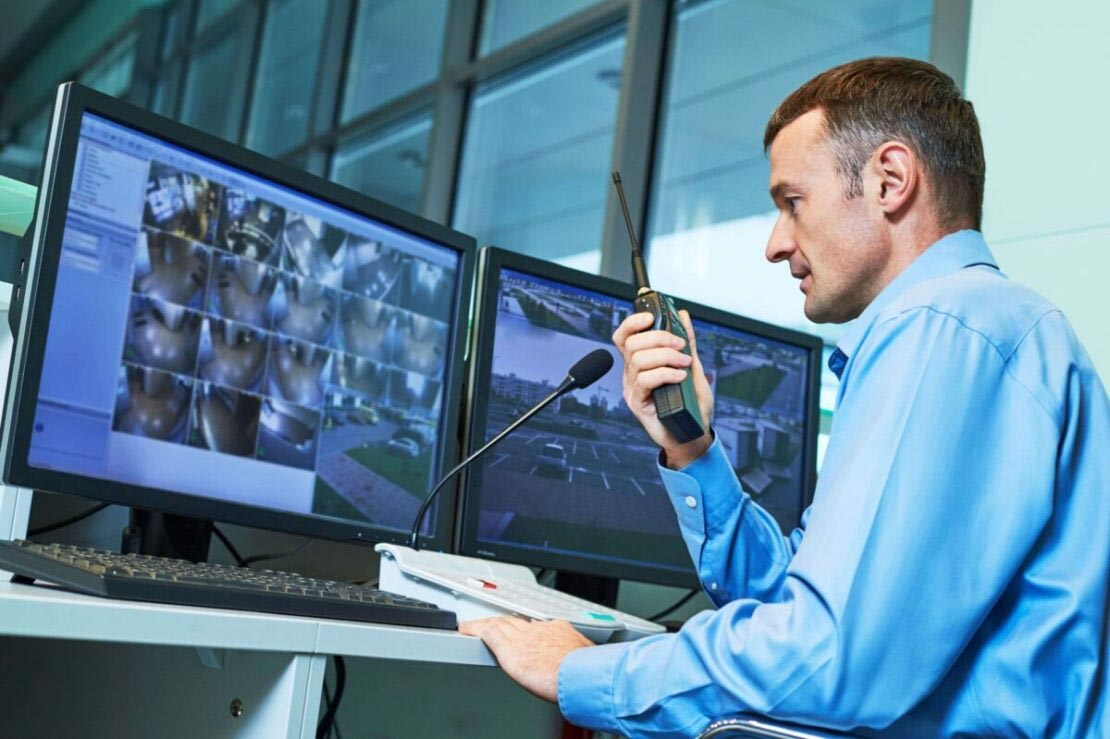 A security guard is talking into a walkie talkie while looking at a computer screen.