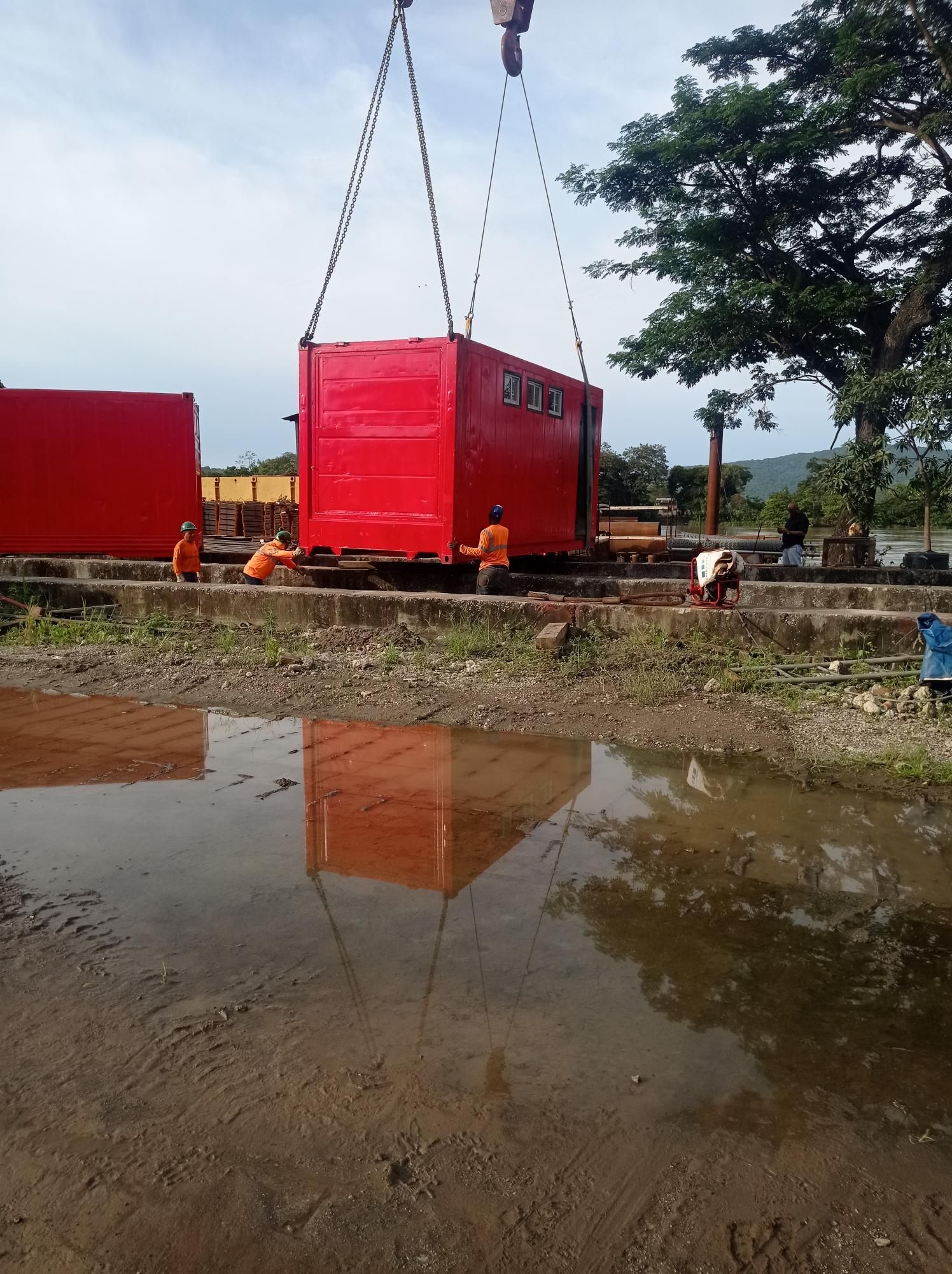 Una grúa baja un gran contenedor rojo mientras unos trabajadores permanecen cerca en una obra al aire libre