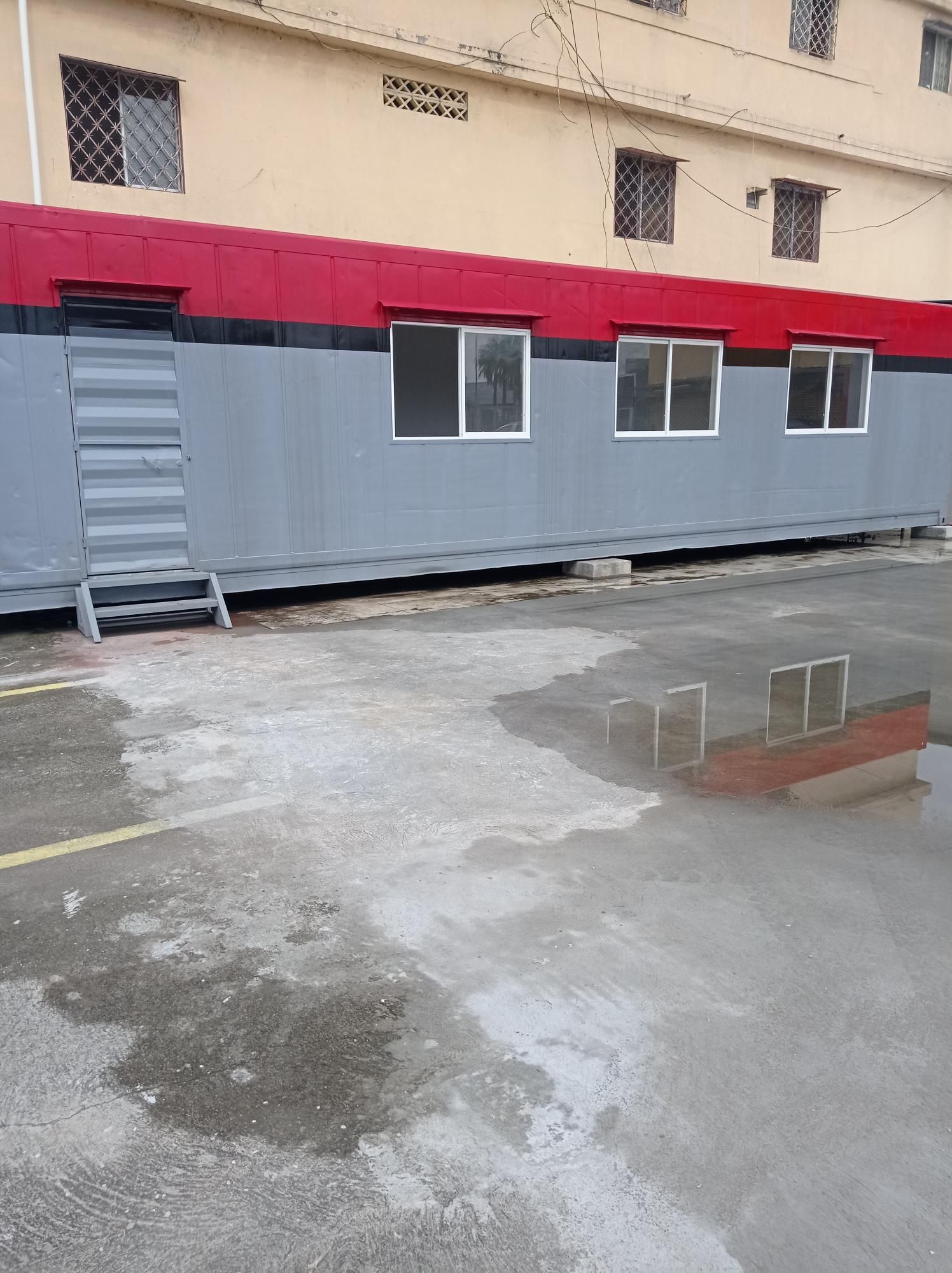 Un edificio modular largo y rectangular, pintado de gris y rojo, situado en una zona pavimentada frente a un edificio beige