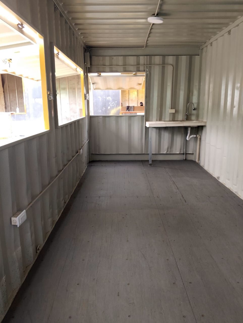 Interior vacío de un contenedor de transporte con paredes y suelo grises, ventanas y un pequeño lavabo junto a la pared.