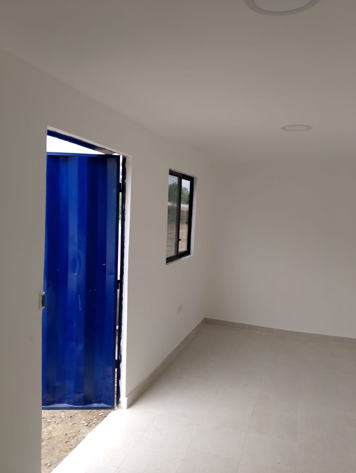 Vista interior de una habitación con paredes y suelo blancos, con una puerta azul oscuro y una pequeña ventana cuadrada.