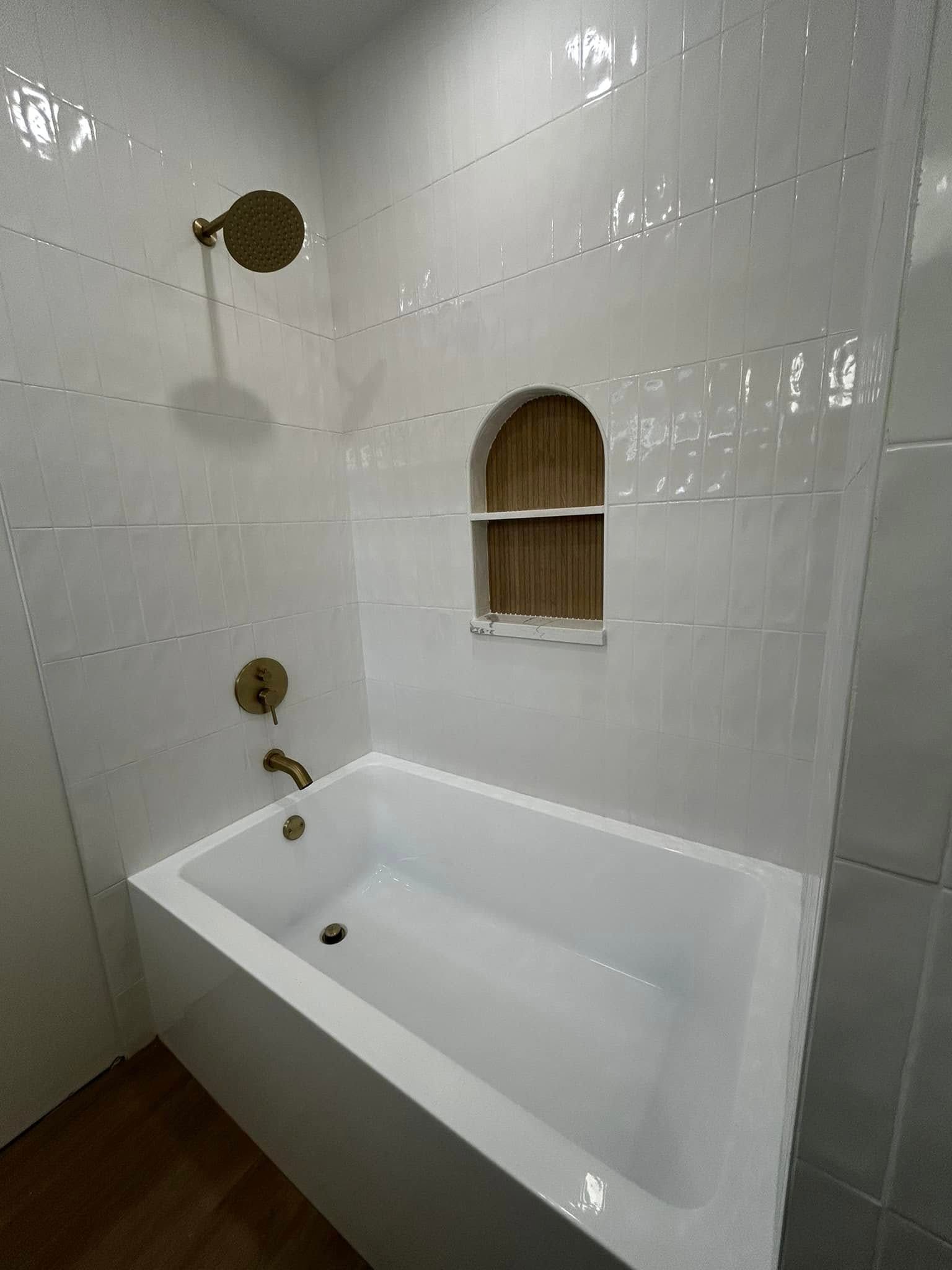 White tiled bathroom with a bathtub, brass shower fixtures, and a small arched niche shelf.