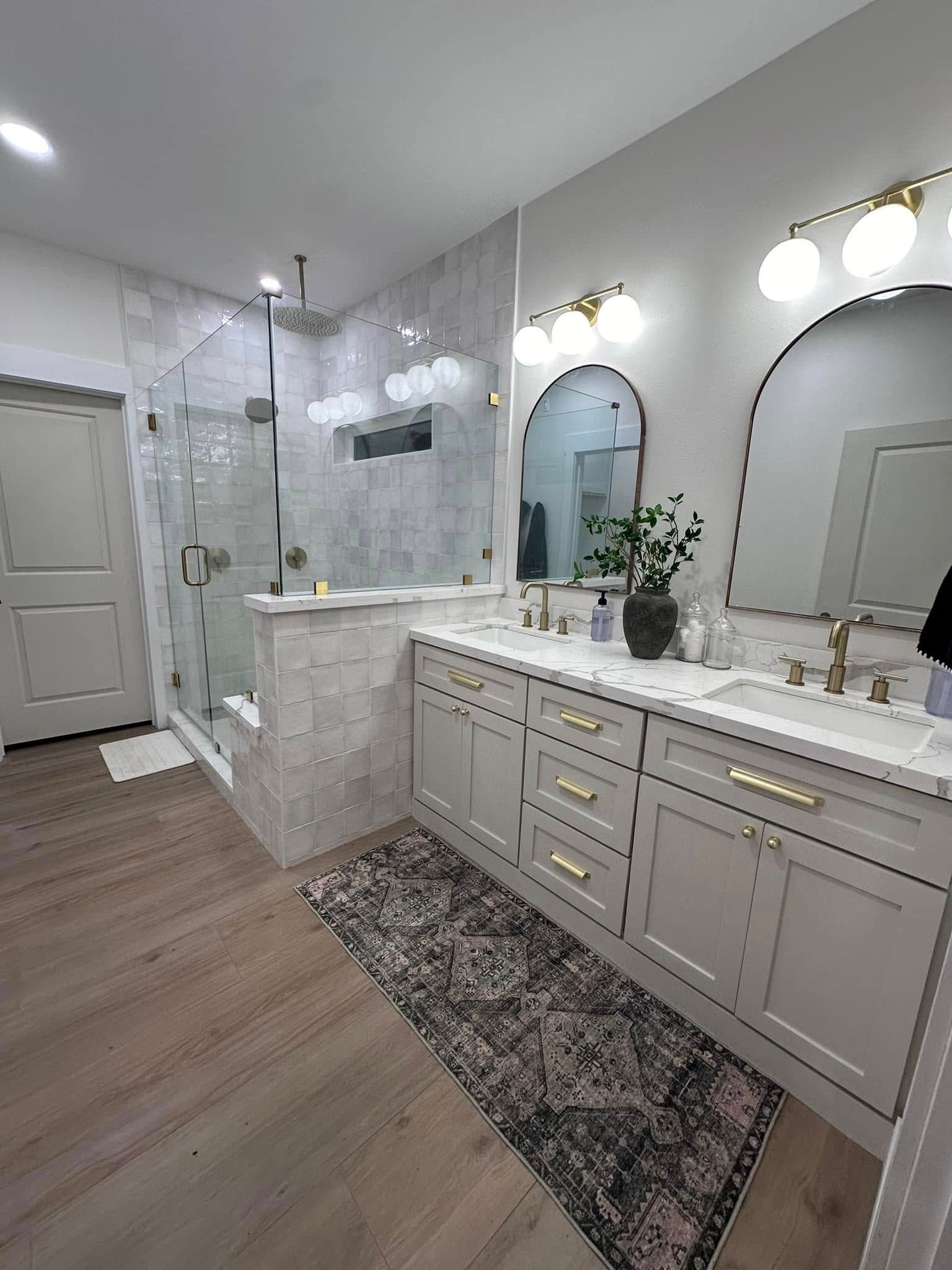 Bright bathroom with double vanity, arched mirrors, glass shower, white cabinets, and patterned rug.