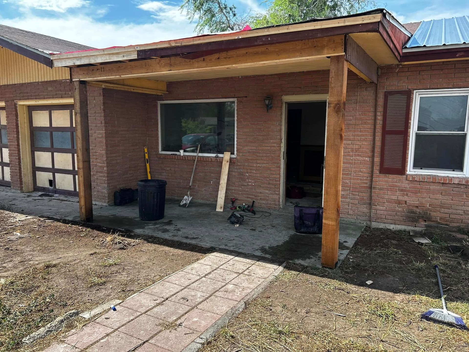 Small brick house porch with wooden posts, open doorway, and a paved walkway in a yard.
