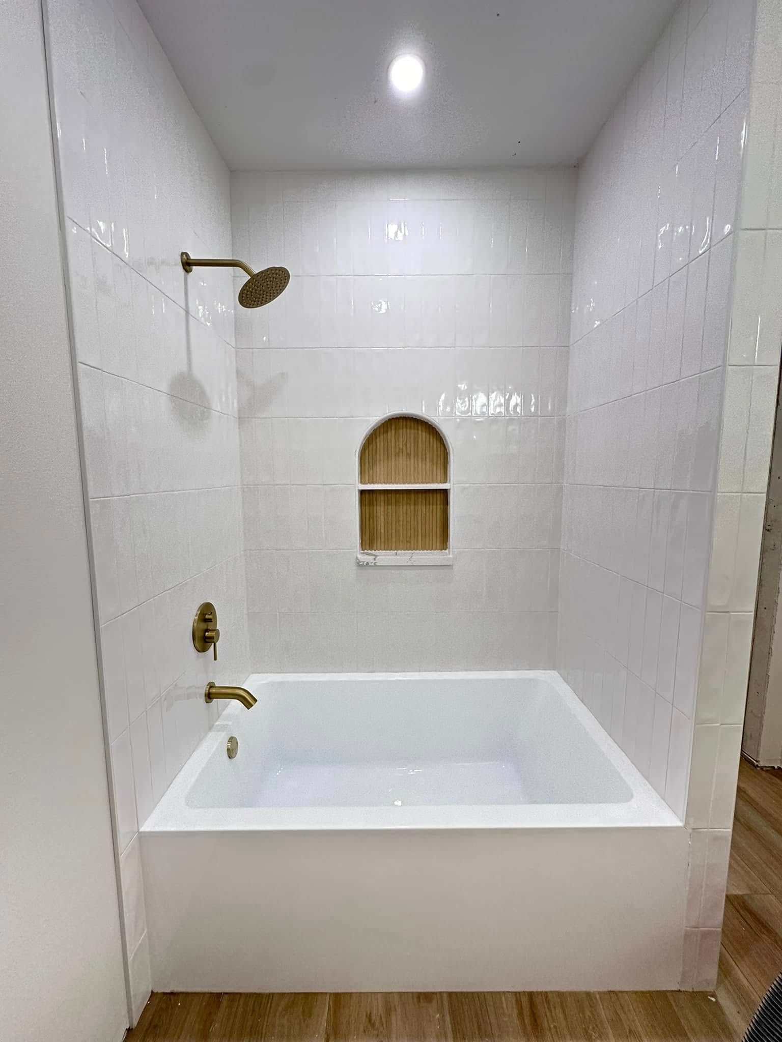 White tiled bathroom with a white bathtub, gold fixtures, and a recessed wall niche.