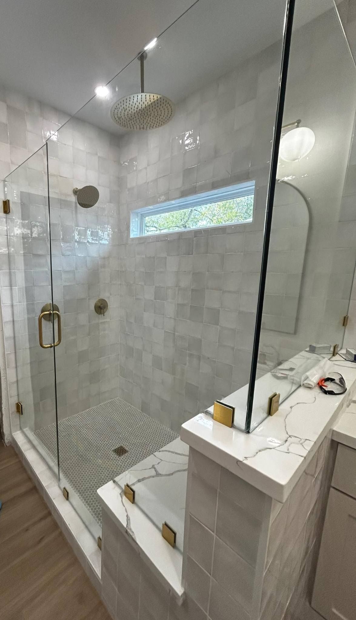Modern bathroom with glass-enclosed shower, white tile, gold fixtures, and a narrow window.