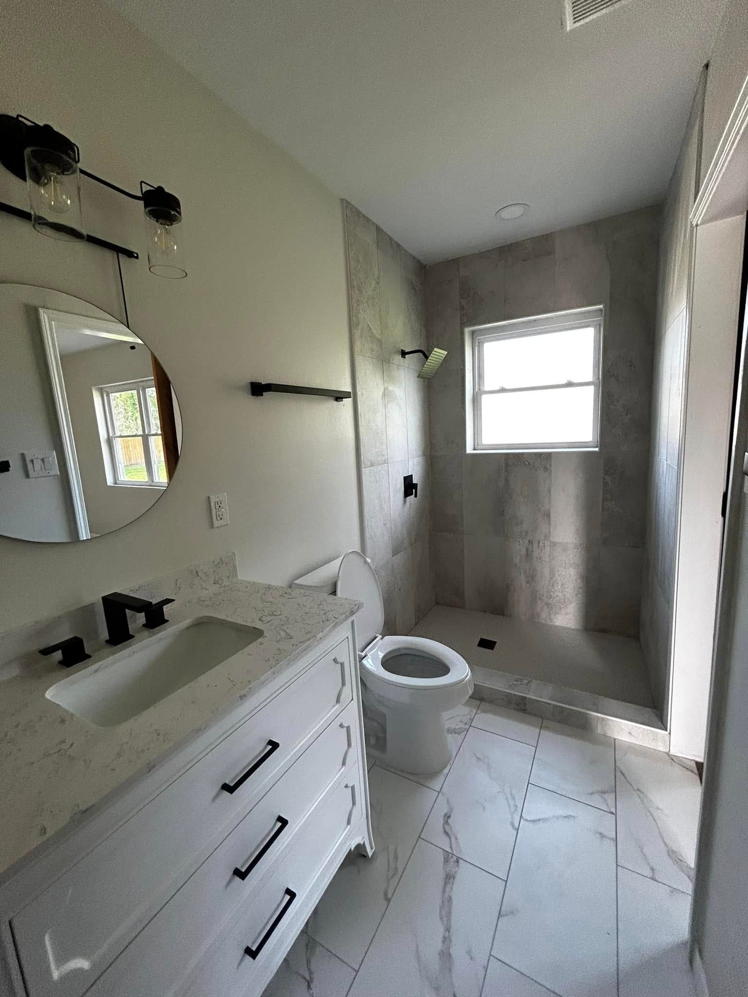 Bright bathroom with white vanity, toilet, tiled shower, and window.