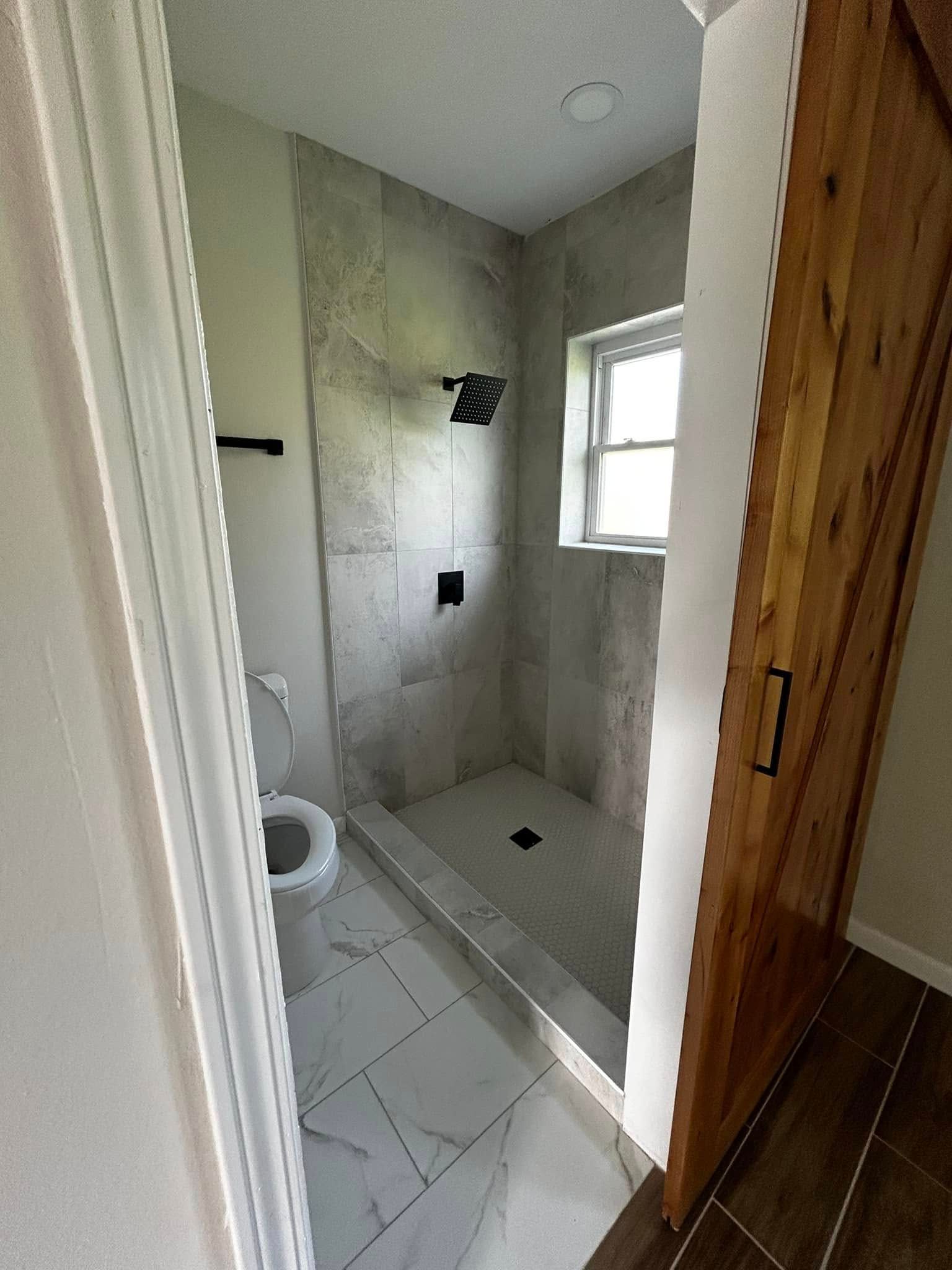 Small tiled bathroom with toilet, shower stall, and window, viewed from the doorway