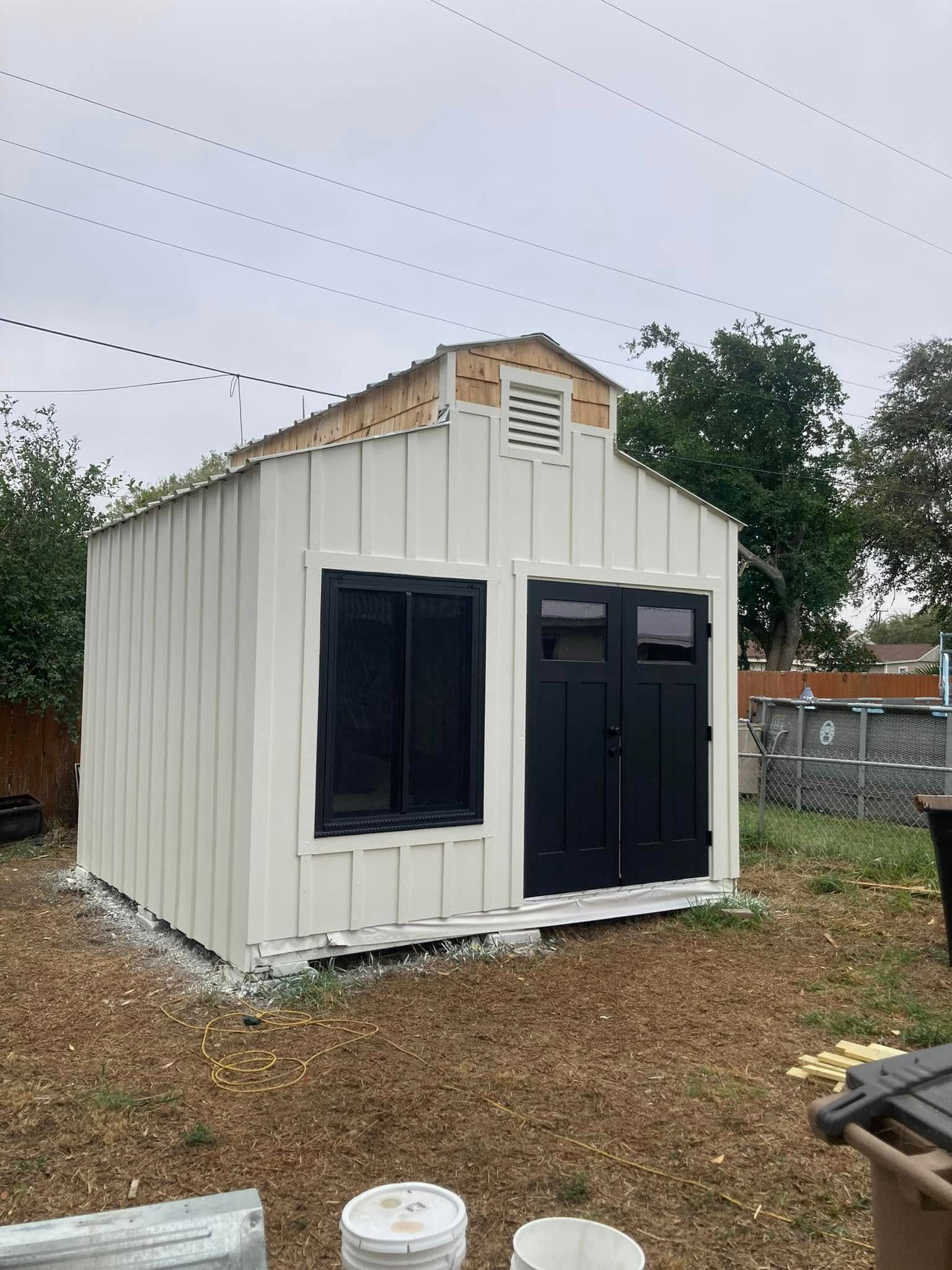 Small white utility shed with dark doors and windows in a yard under an overcast sky.