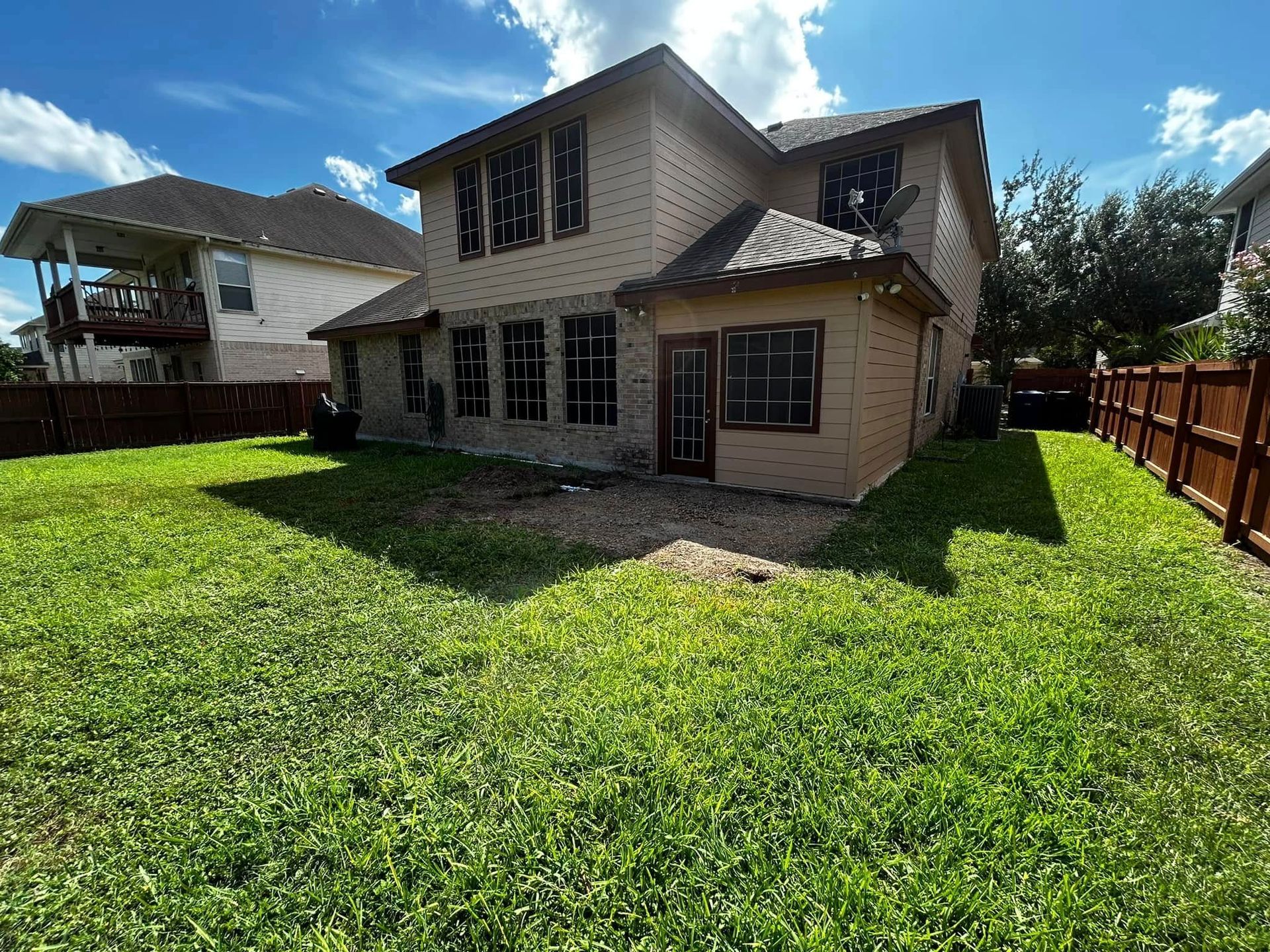 Two-story brick house with a large green backyard and wooden fence under a sunny blue sky