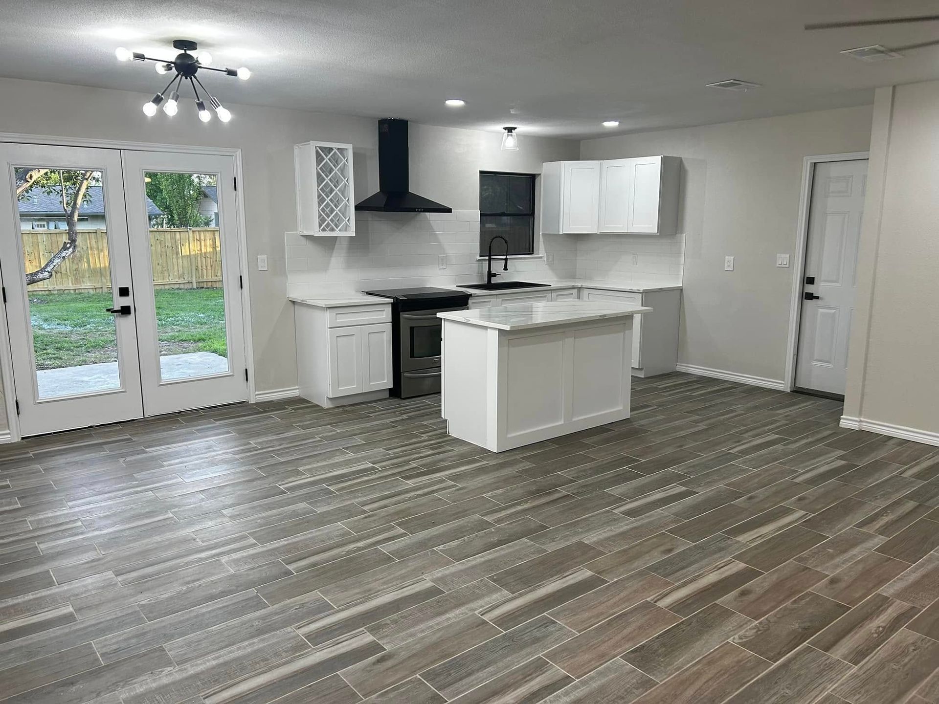 Empty modern kitchen and dining area with white cabinets, island, black range hood, and French doors to yard