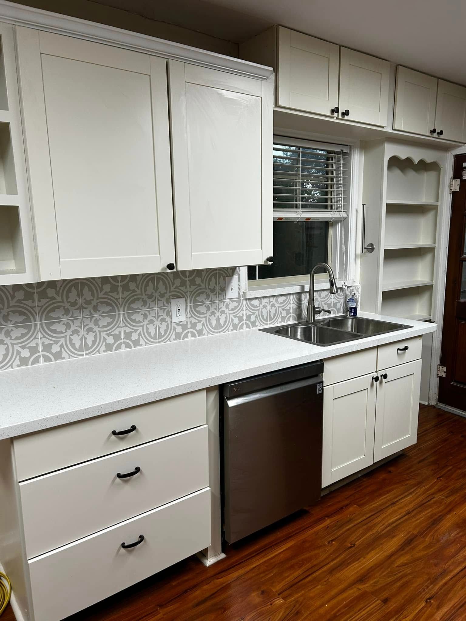 White kitchen with cabinets, patterned backsplash, sink under window, dishwasher, and wood floor