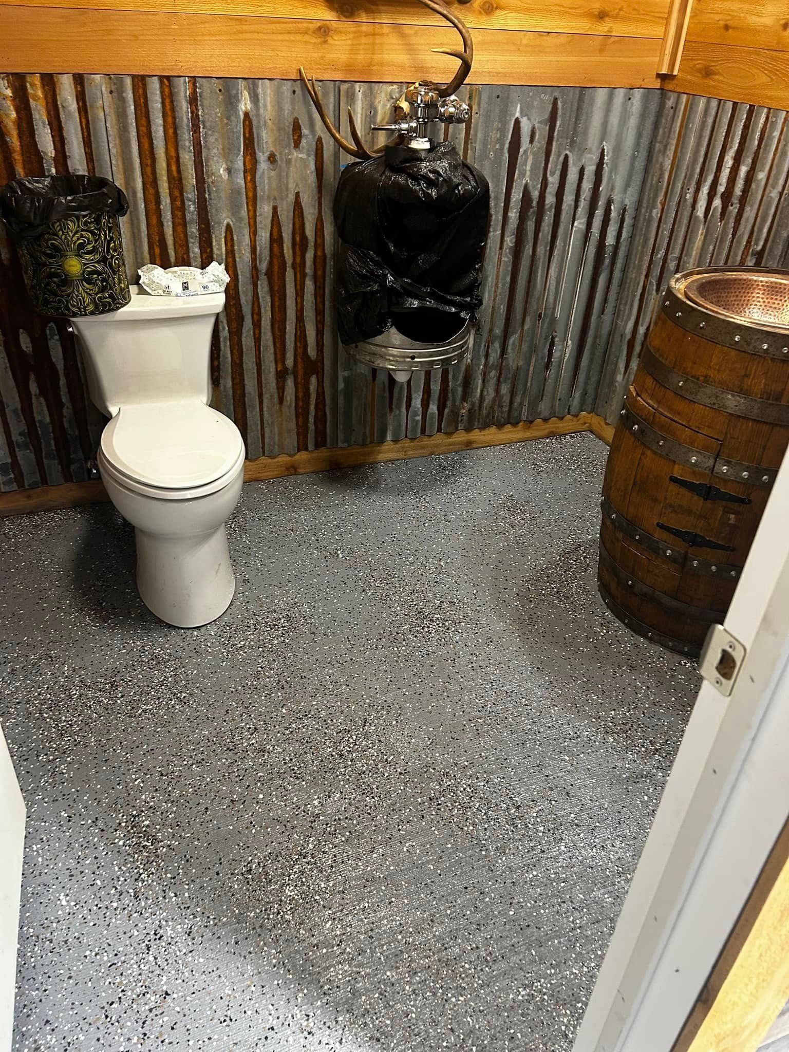 Rustic outdoor restroom with a white toilet, corrugated metal walls, and a speckled concrete floor.