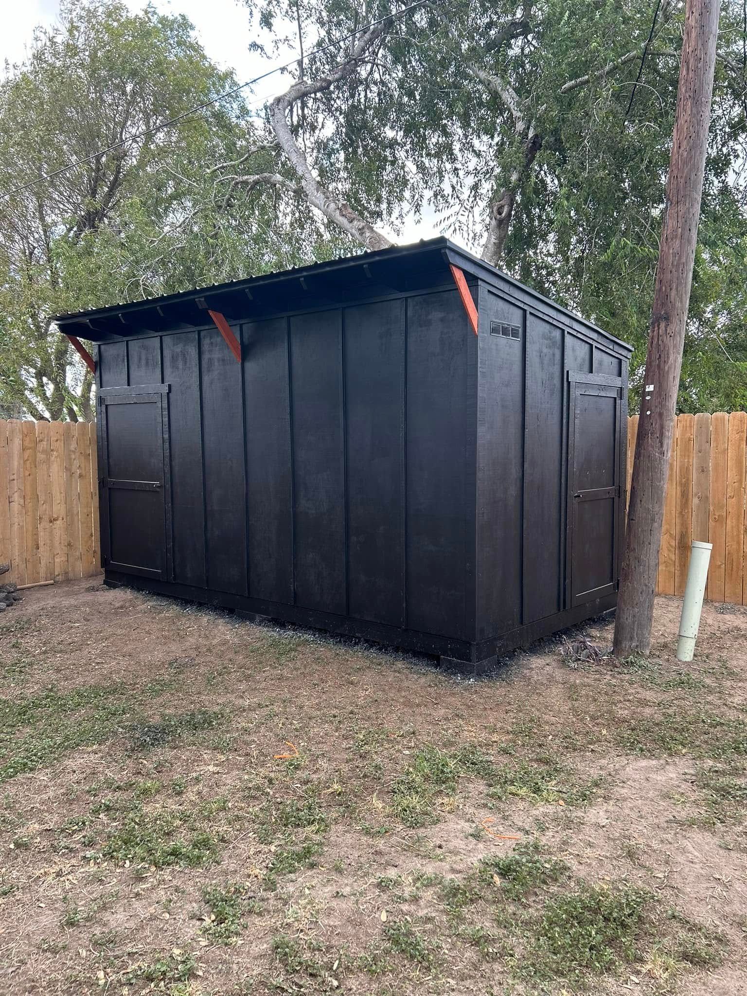 Black storage shed with a dark roof in a fenced backyard, surrounded by trees and gravel.