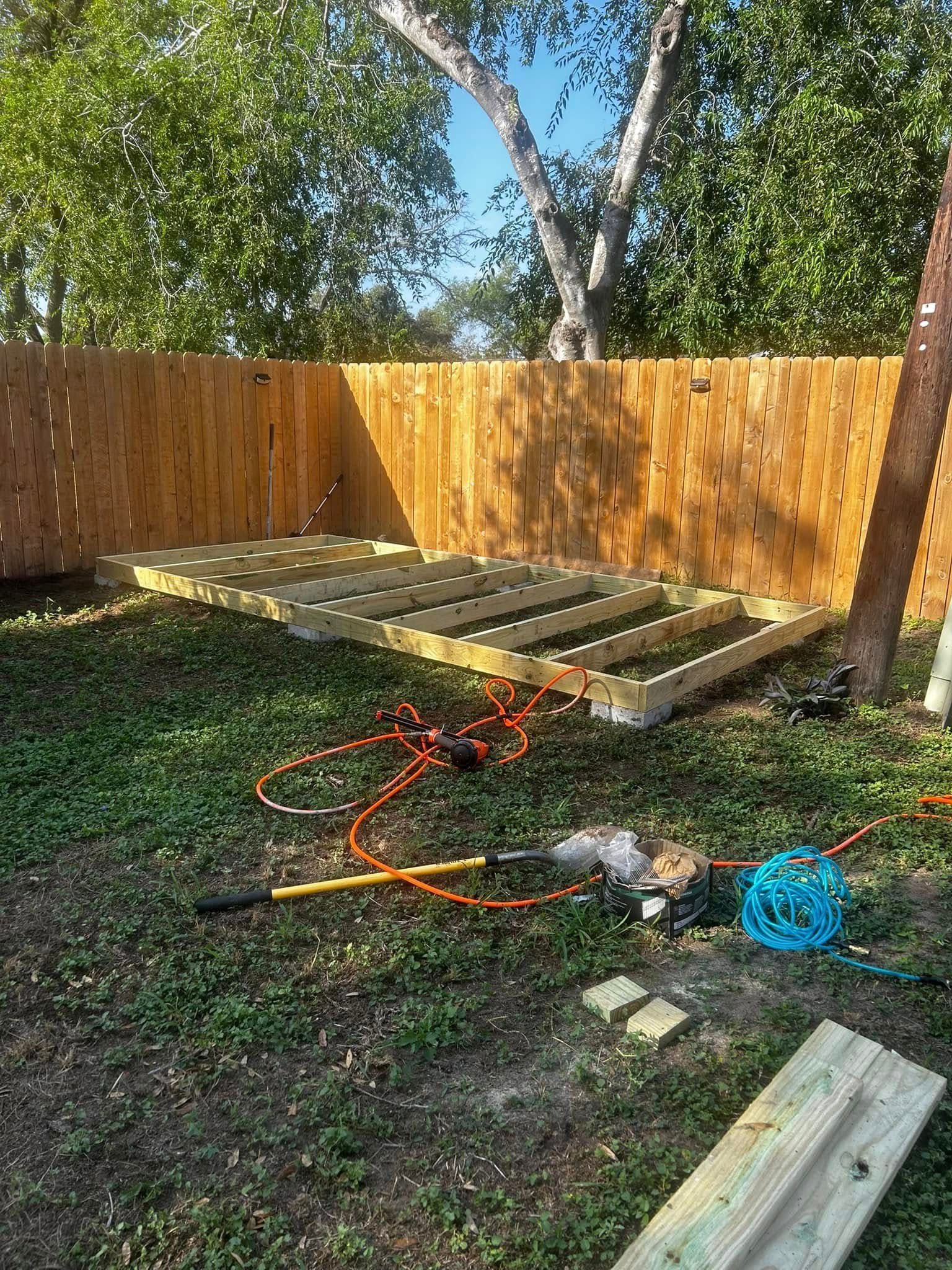 Backyard with a wooden fence, raised deck frame, tools, and scattered lumber under trees