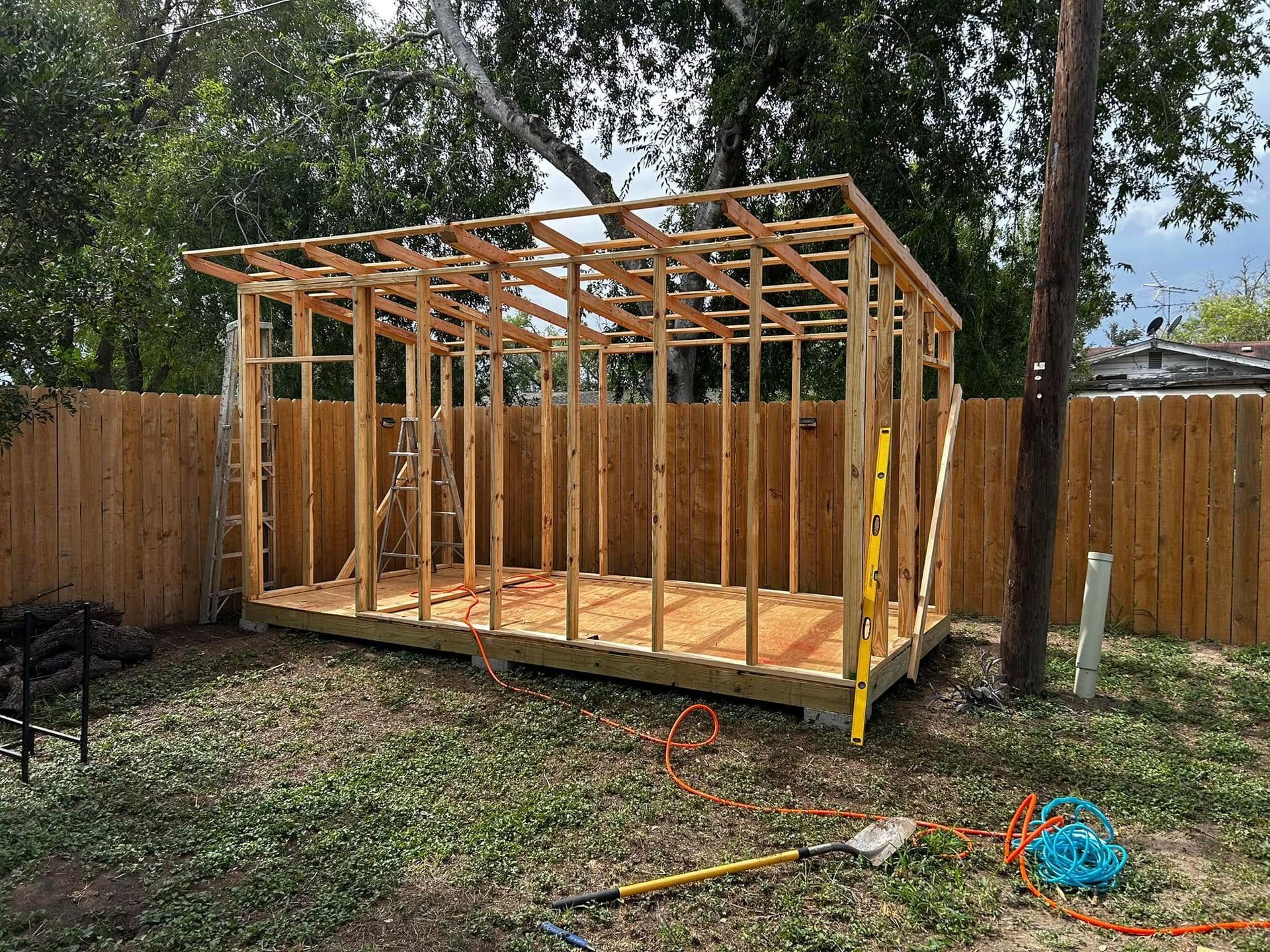 Partially built wooden shed frame in a fenced backyard, with tools on the grass.