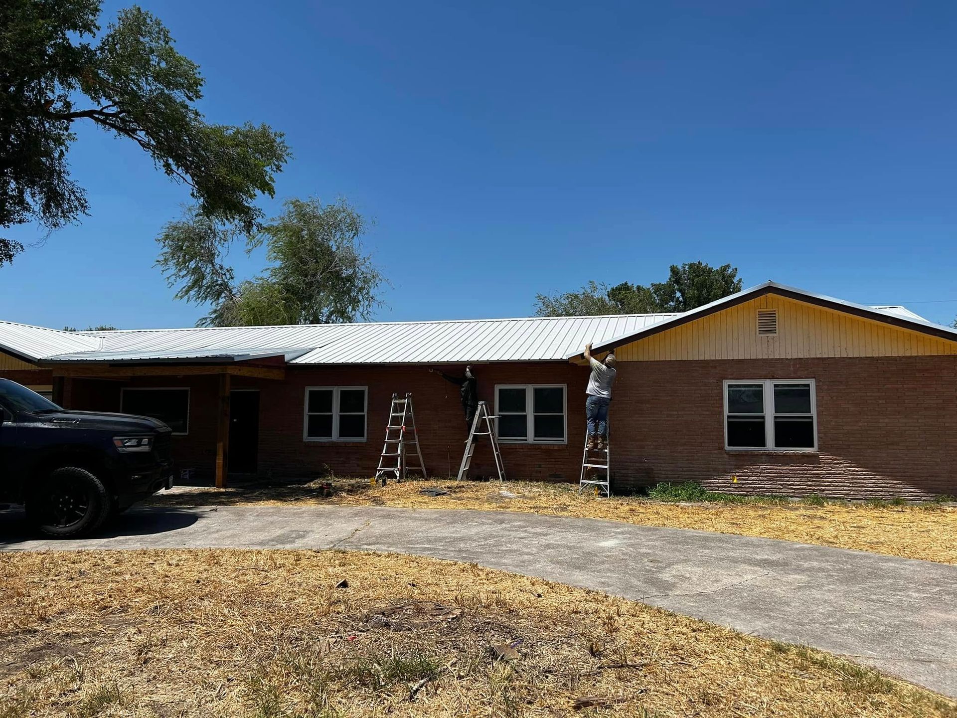Single-story brick house with a metal roof, ladder on the porch, and a driveway in front.