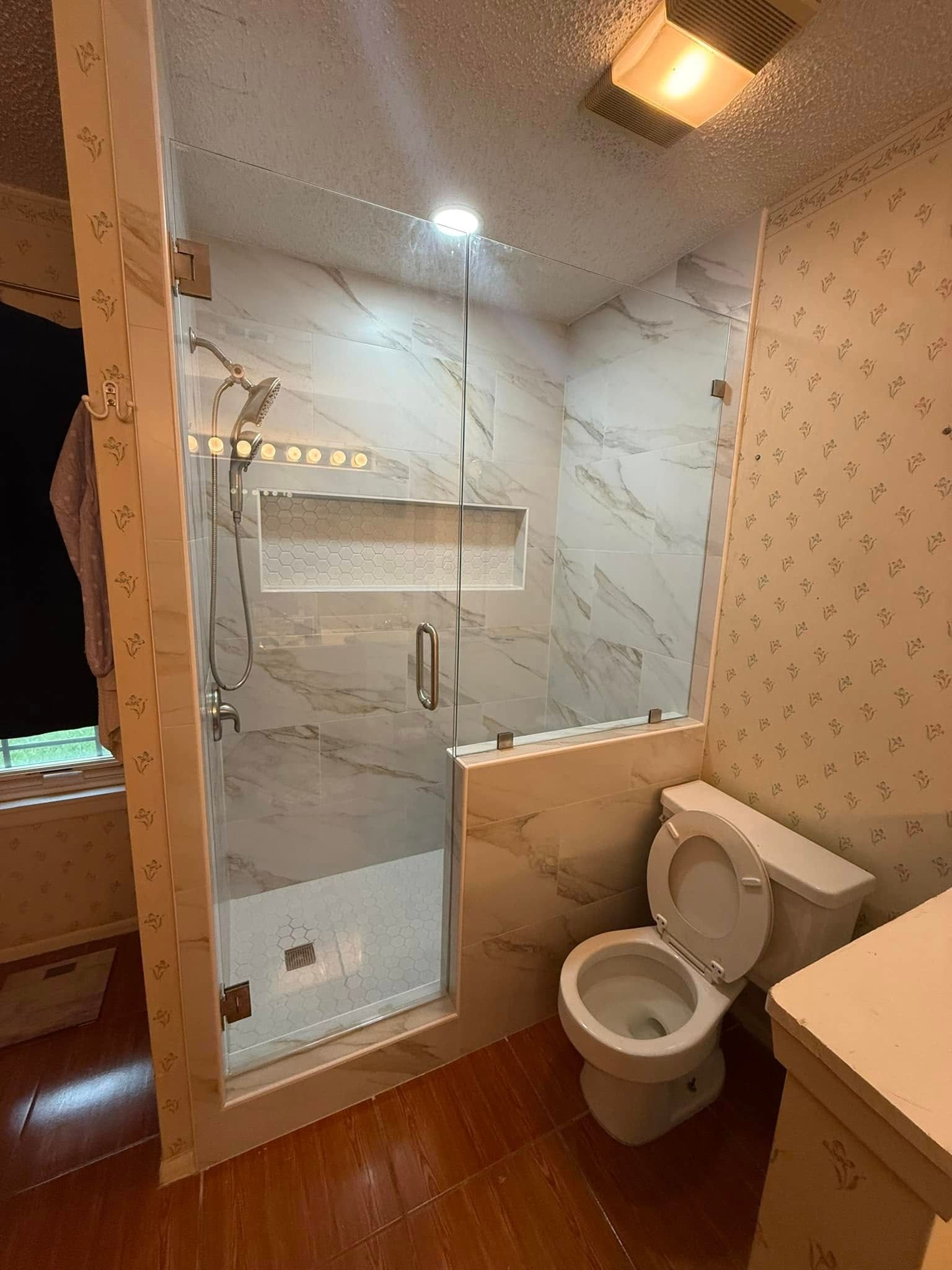 Compact bathroom with white marble tiles, glass shower, toilet, and wooden floor-lit by warm ceiling lights