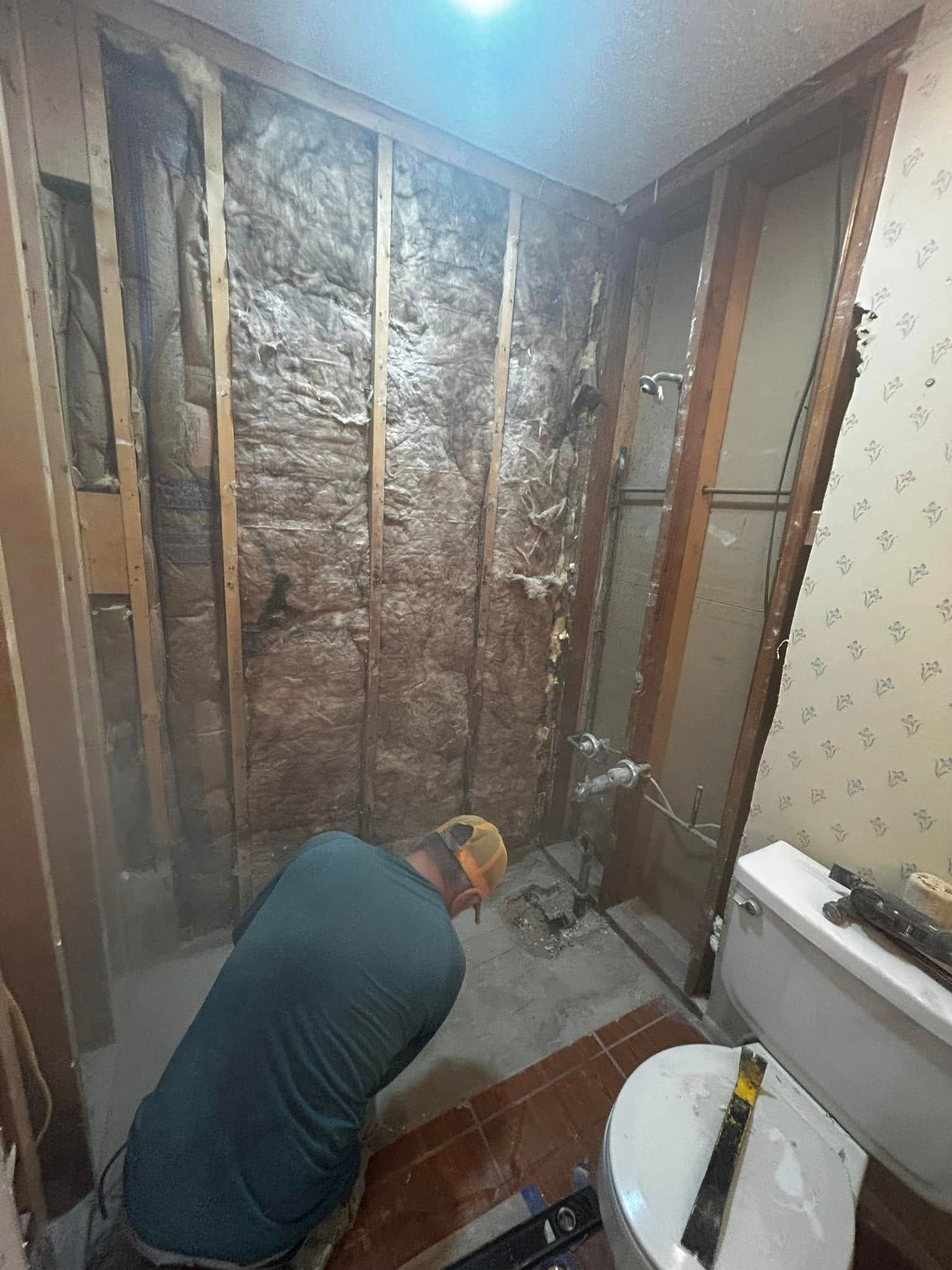 Person kneels in a bathroom under renovation, exposed studs and insulation visible on the walls.