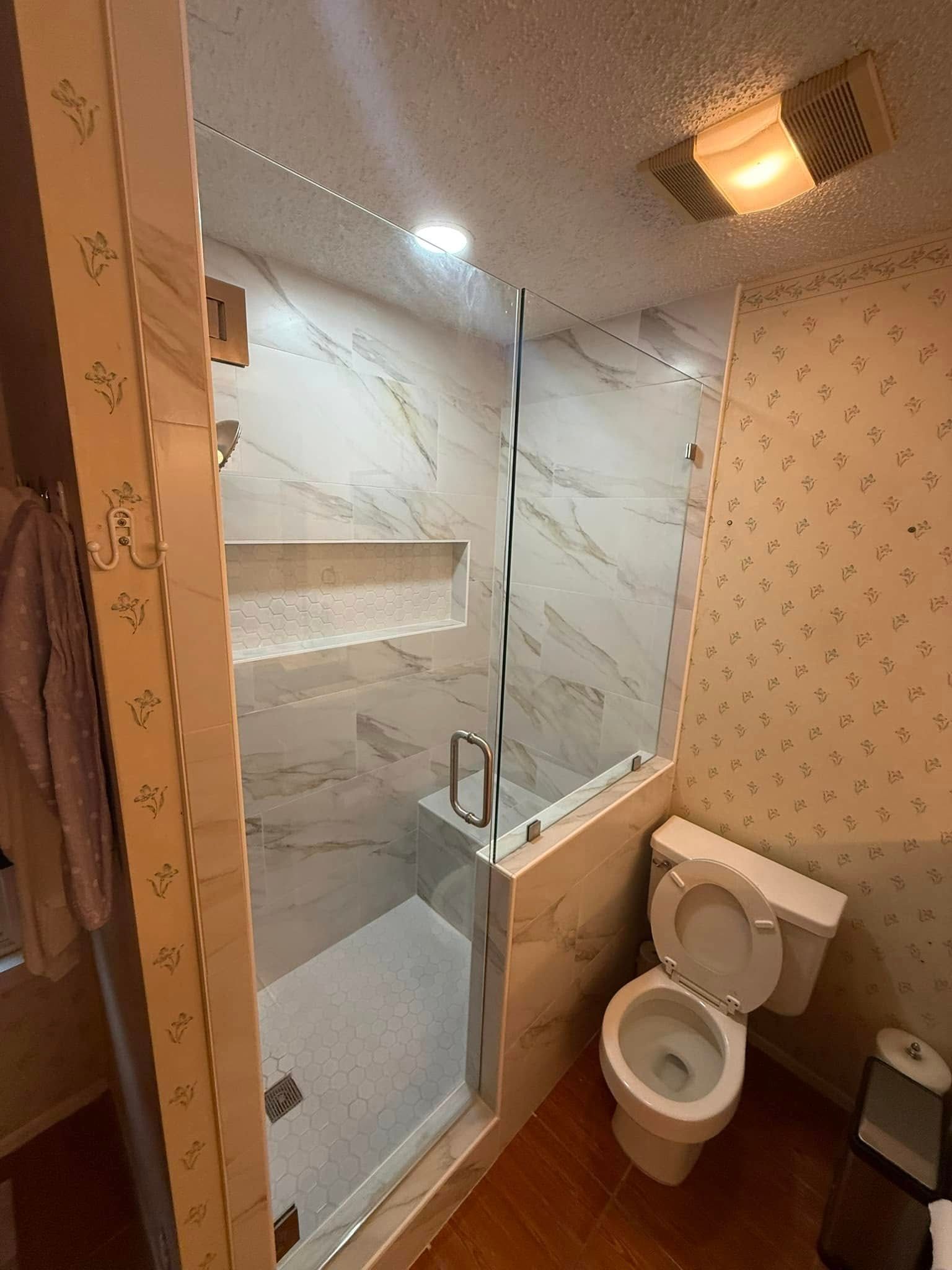 Small bathroom with glass shower enclosure, white marble walls, toilet, and warm ceiling light.