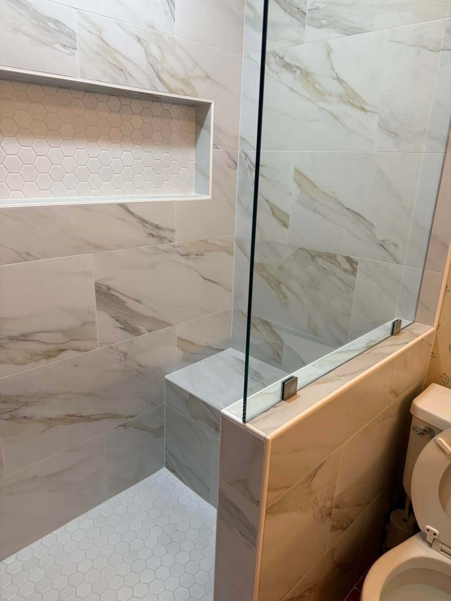 Small bathroom shower with beige tile, glass partition, recessed shelf, and toilet visible on the right.