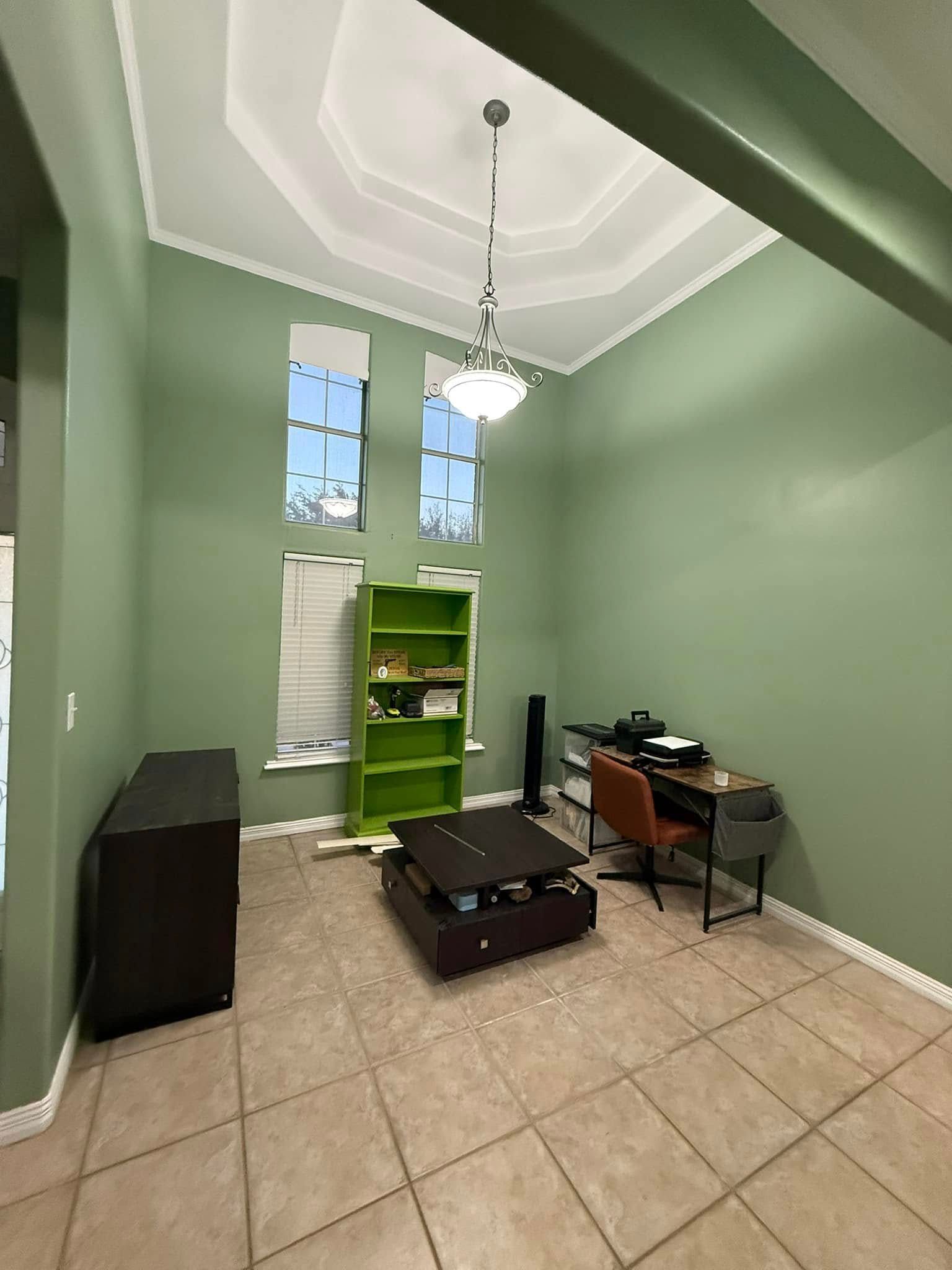 Small green room with tiled floor, desk, chair, suitcase, and hanging light beneath tall windows.