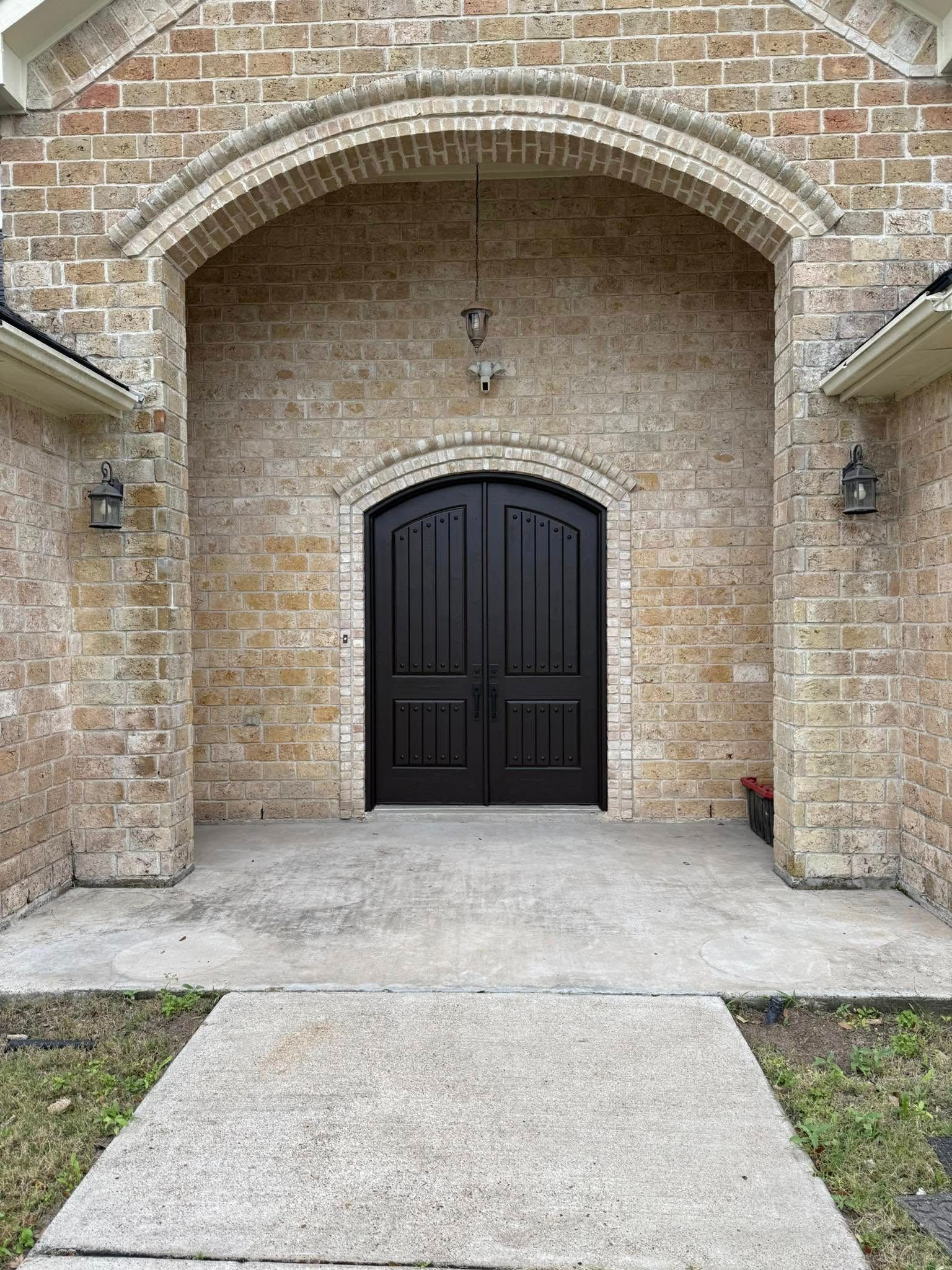 Brick front entry with arched alcove and double black doors, framed by concrete walkway and grass.