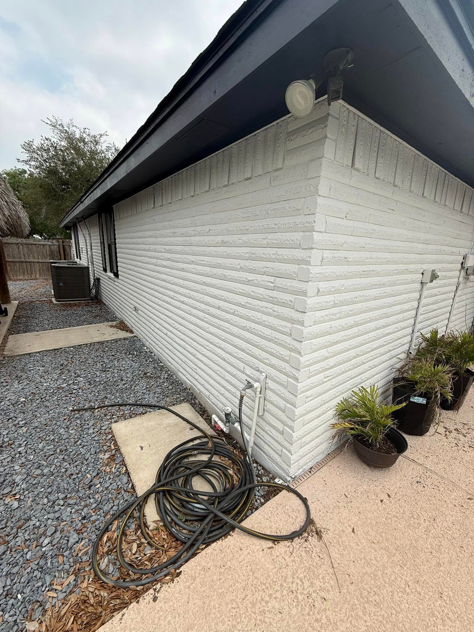 Side of white brick house with black roof, gravel path, coiled hose, and potted plants by the wall.